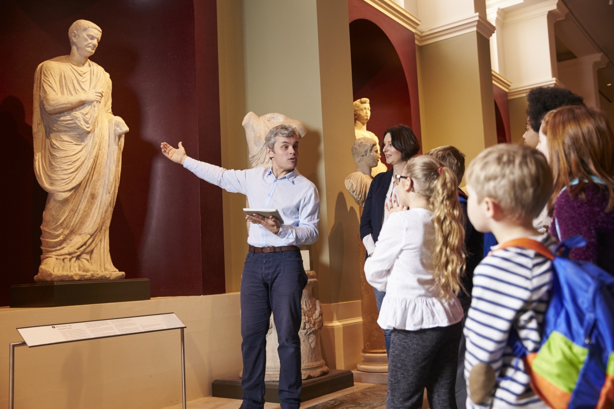 man gesturing to a statue while giving a presentation to a group of children in a gallery or museum