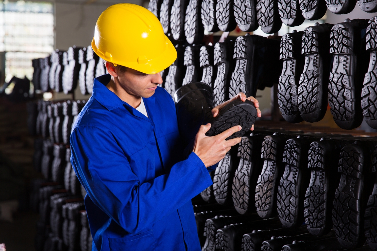 person, wearing a hard hat, inspecting the soles of newly-manufactured safety shoes