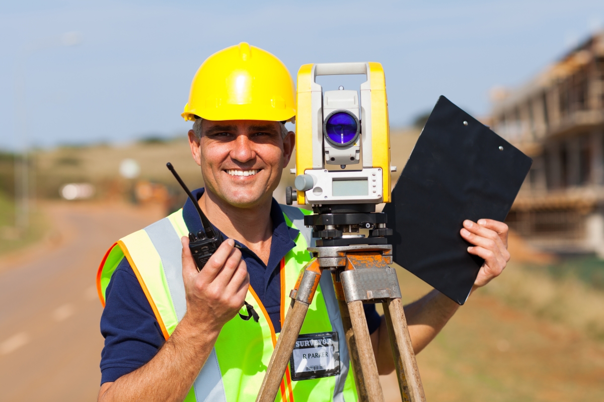 man in safety vest and hard hat holding a walkie talkie and clipboard standing next to a land surveying instrument on a tripod