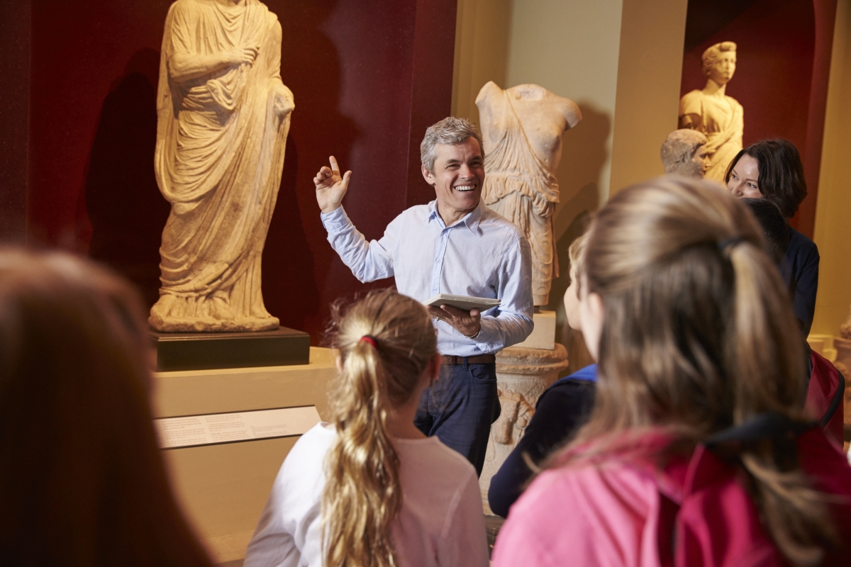 man pointing at statues in front of a group of people in a gallery or museum