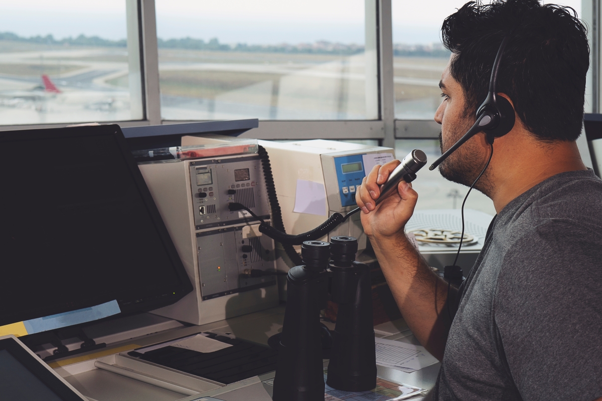bipoc male wearing a headset and speaking into a microphone sitting at a desk with equipment in front of him, an airport is visible outside the window
