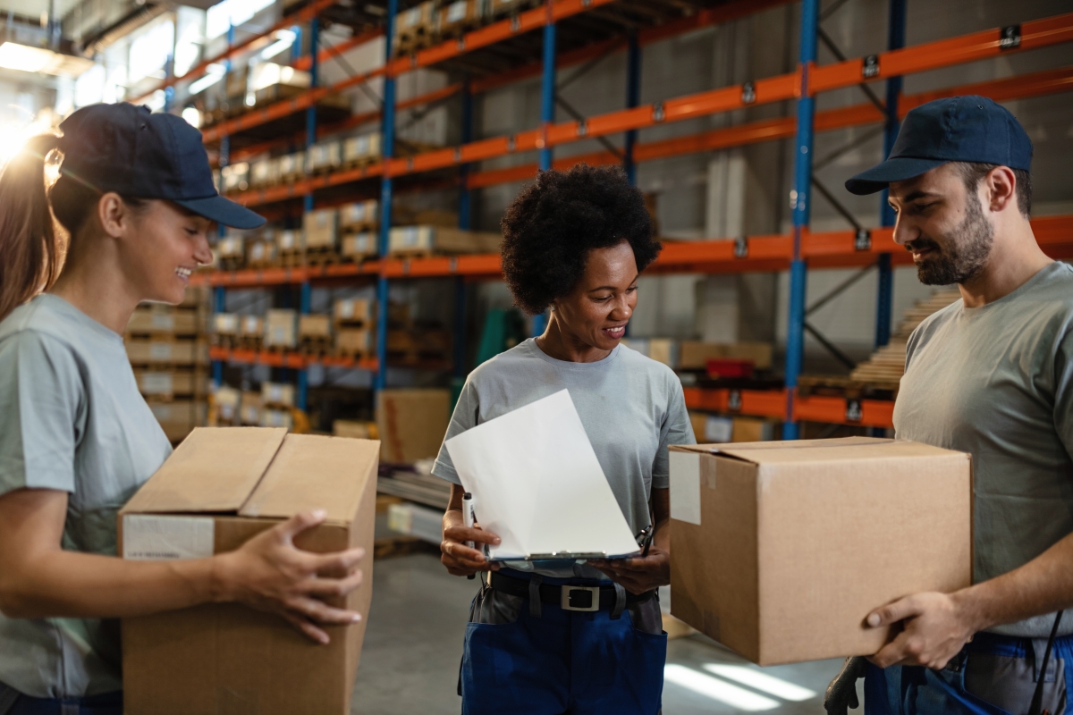 three diverse people, two holding boxes and one holding a pen and clipboard, standing in a warehouse