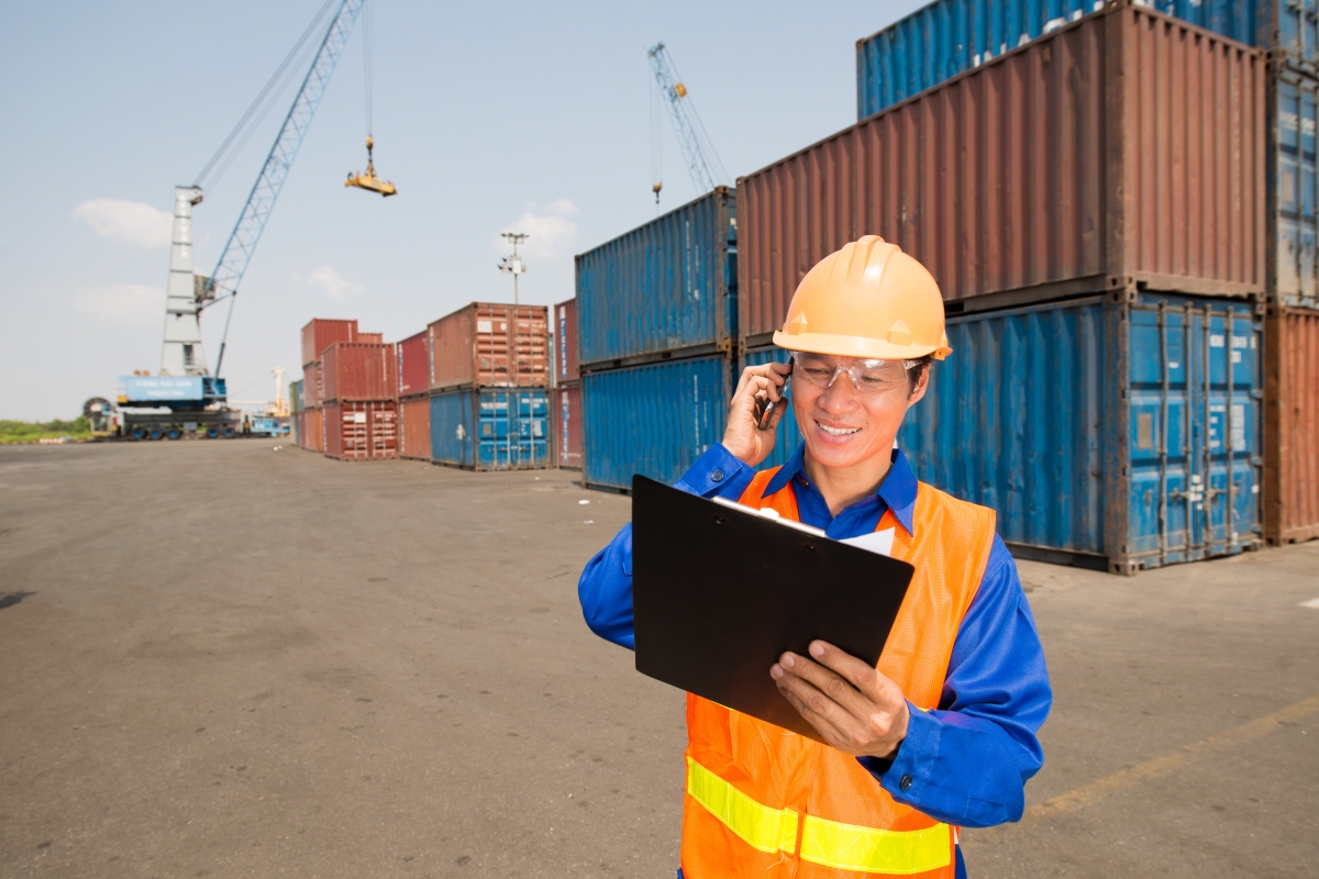 man in hard hat, safety glasses and safety vest standing near many cargo containers talking on a cellphone while looking at a clipboard