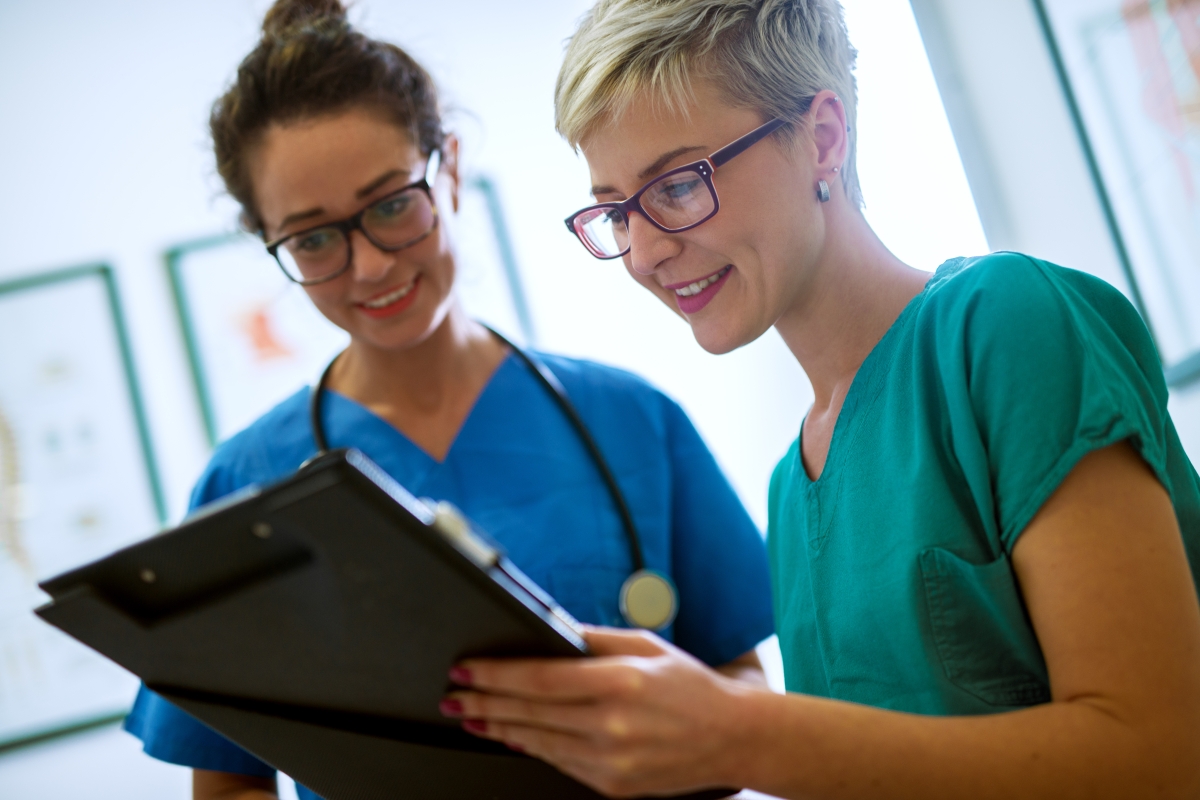 two women wearing scrubs and glasses, one with a stethoscope around her neck, both looking at a clipboard