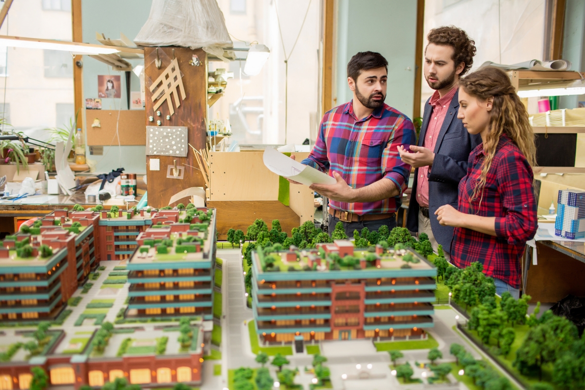 three people standing and talking next to model building and neighborhood plans while one person holds a document
