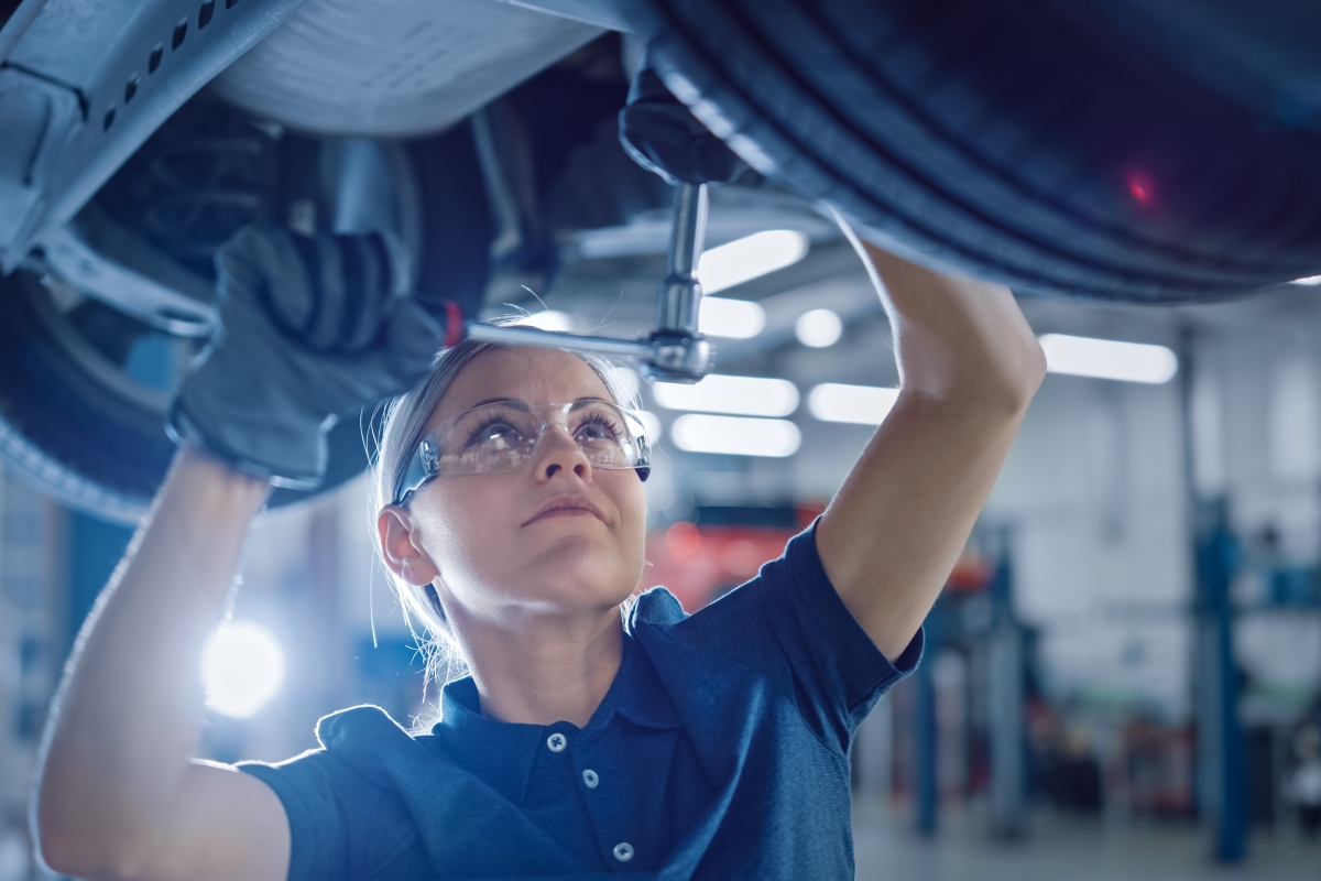 woman wearing safety glasses and gloves working on the underside of a vehicle hoisted in the air in a shop