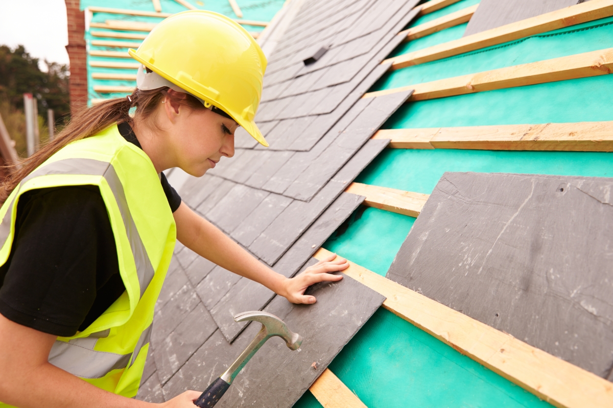 female in hard hat and safety vest using a hammer to tile a roof