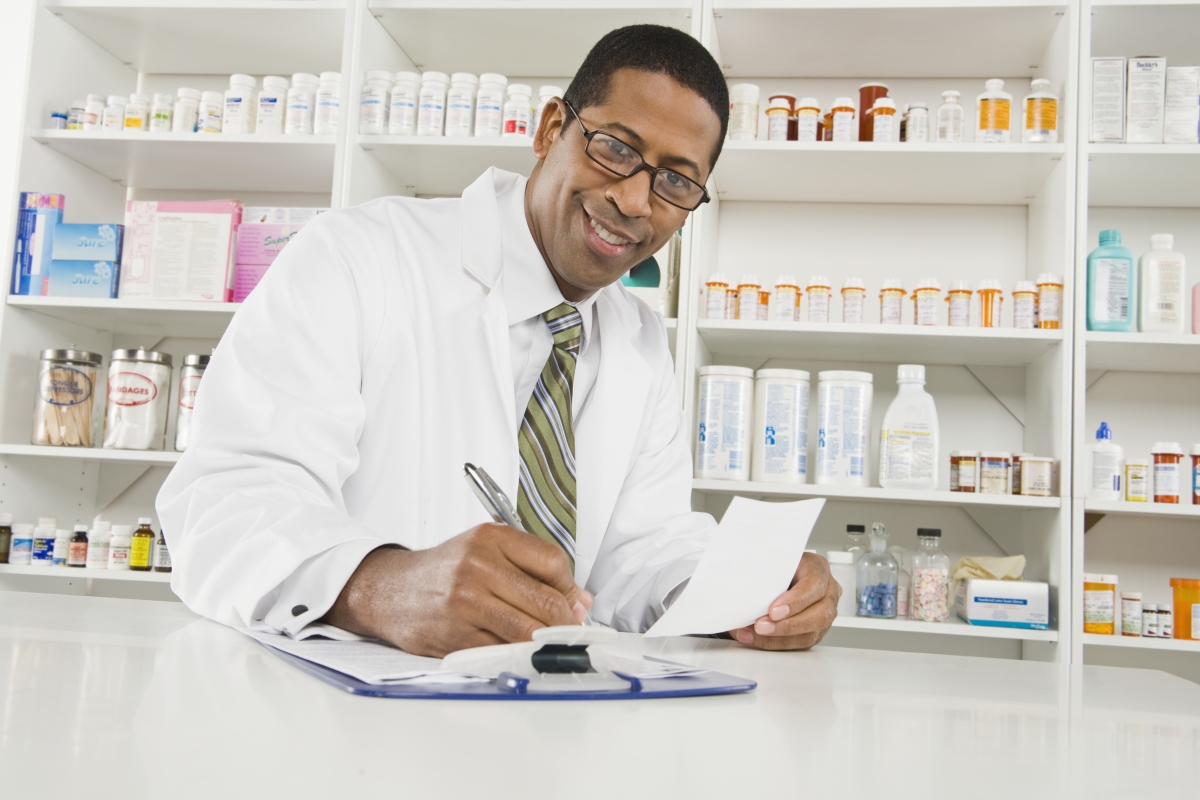 bipoc male pharmacist in a pharmacy holding a prescription in one hand and writing notes on a clipboard with his other hand