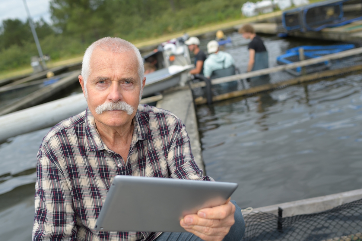 elderly man in a plaid shirt holding a tablet while seated in a fishing boat with another fishing boat and men standing in shallow water in the background