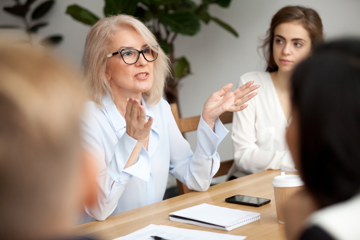 woman with a notepad and phone on the table in front of her speaking to other people all seated around her at the table