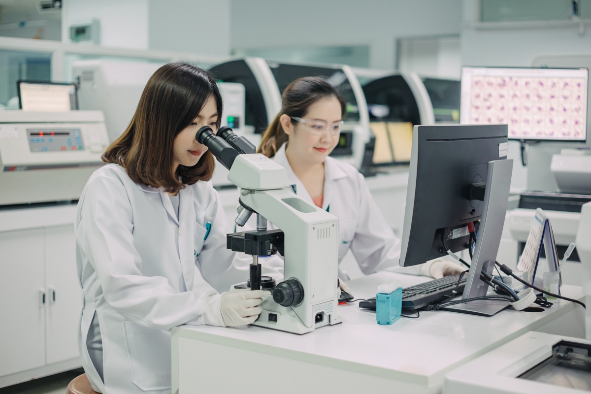 two young bipoc females wearing lab coats in a medical lab while one looks through a microscope and the other works on a computer 