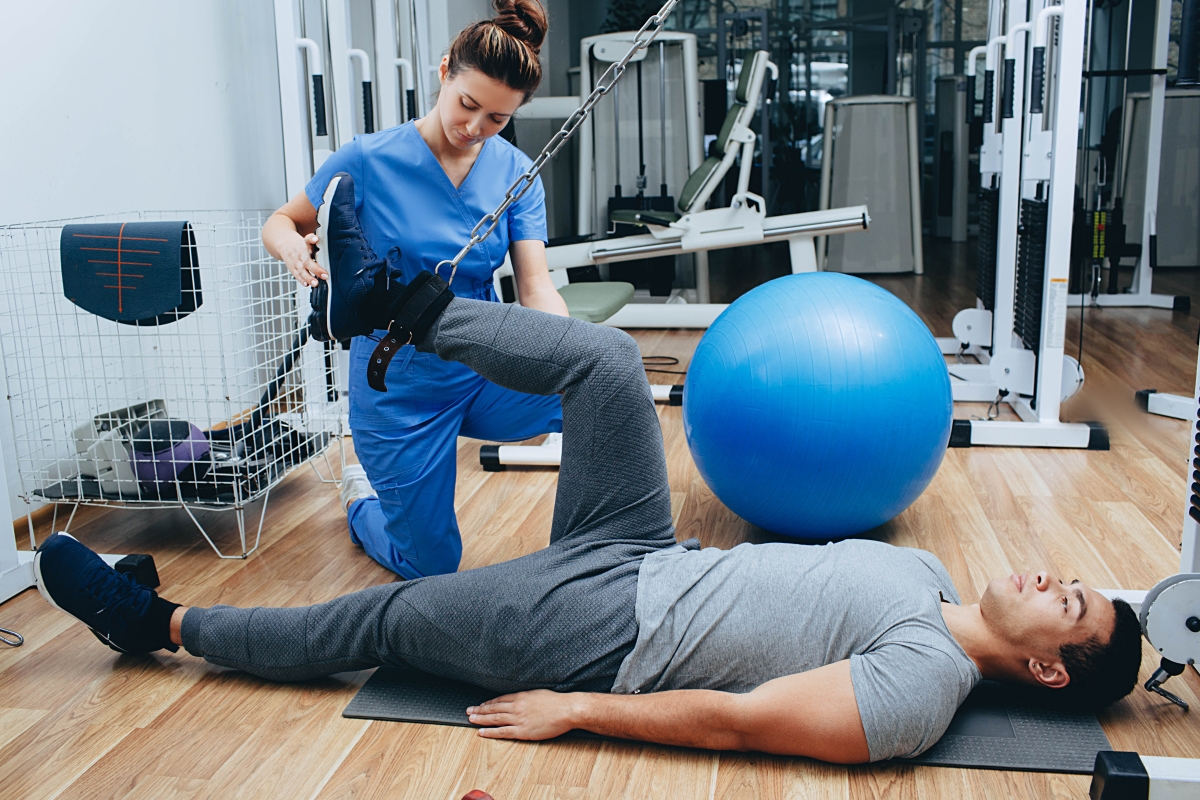 young woman in scrubs lifting and stretching a man's leg while he lies down on a mat in a gym