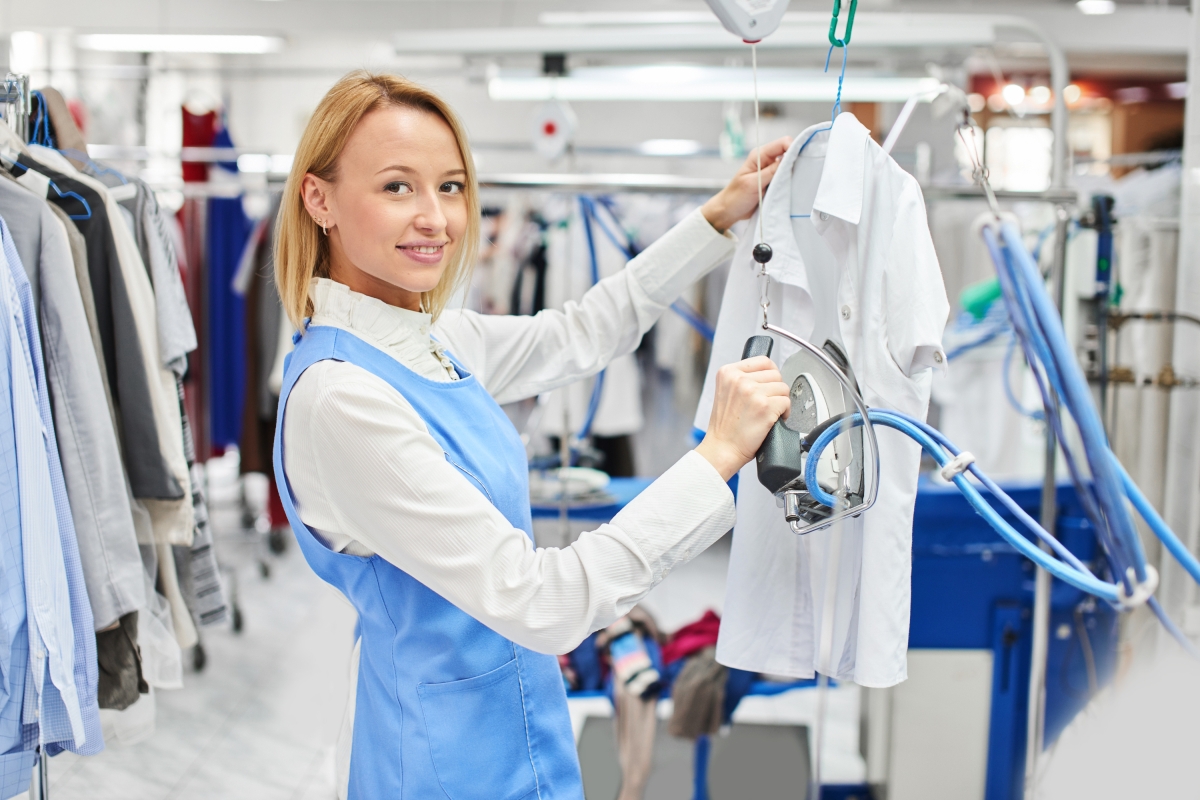 woman in uniform ironing a shirt on a hanger surrounded by other clothes on hangers