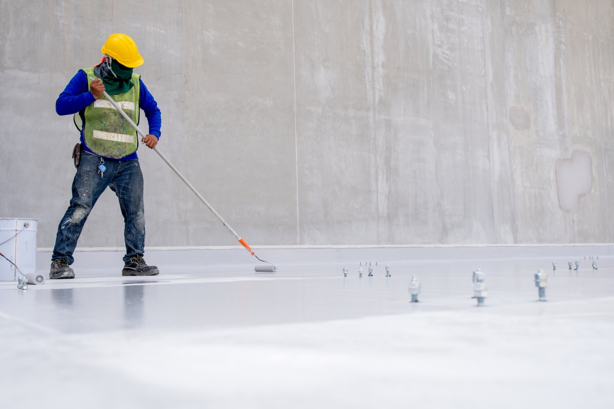 person, wearing a hard hat, safety vest and industrial mask, putting a coating substance on a floor in an industrial setting