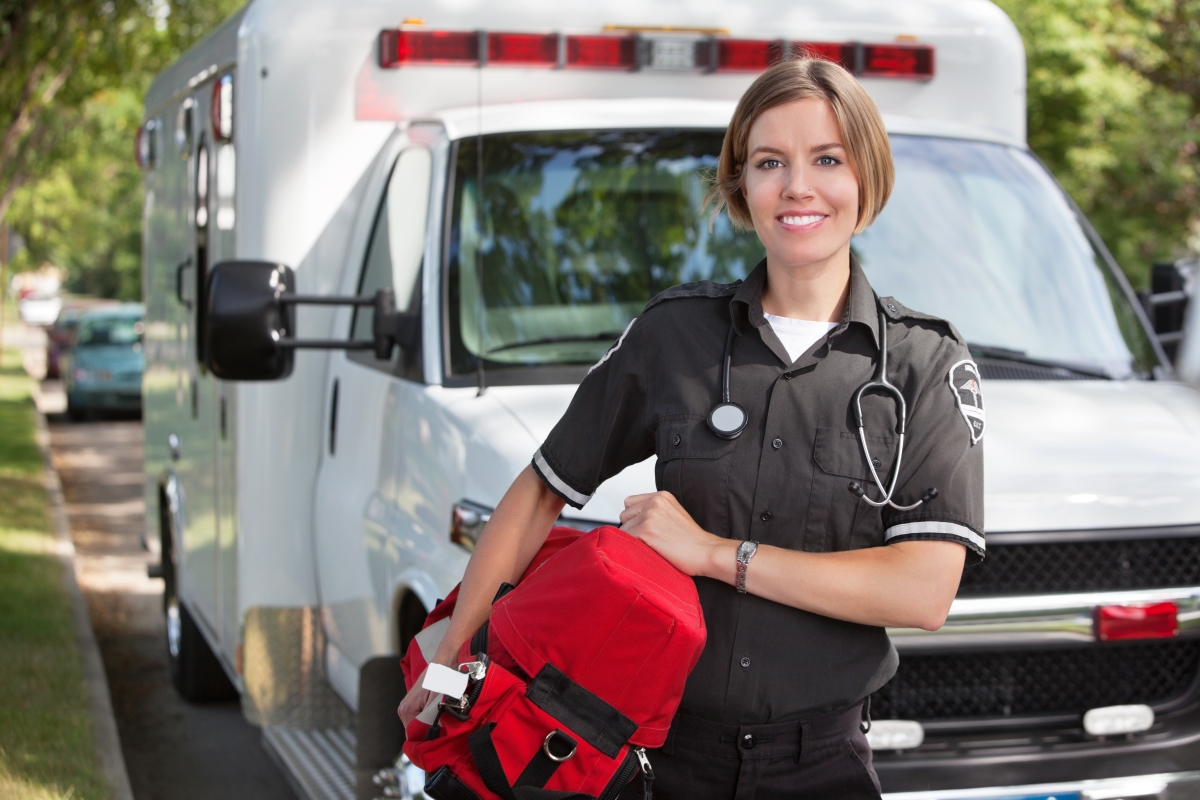 young woman wearing a uniform and stethoscope, holding a red medical bag, standing in front of an ambulance