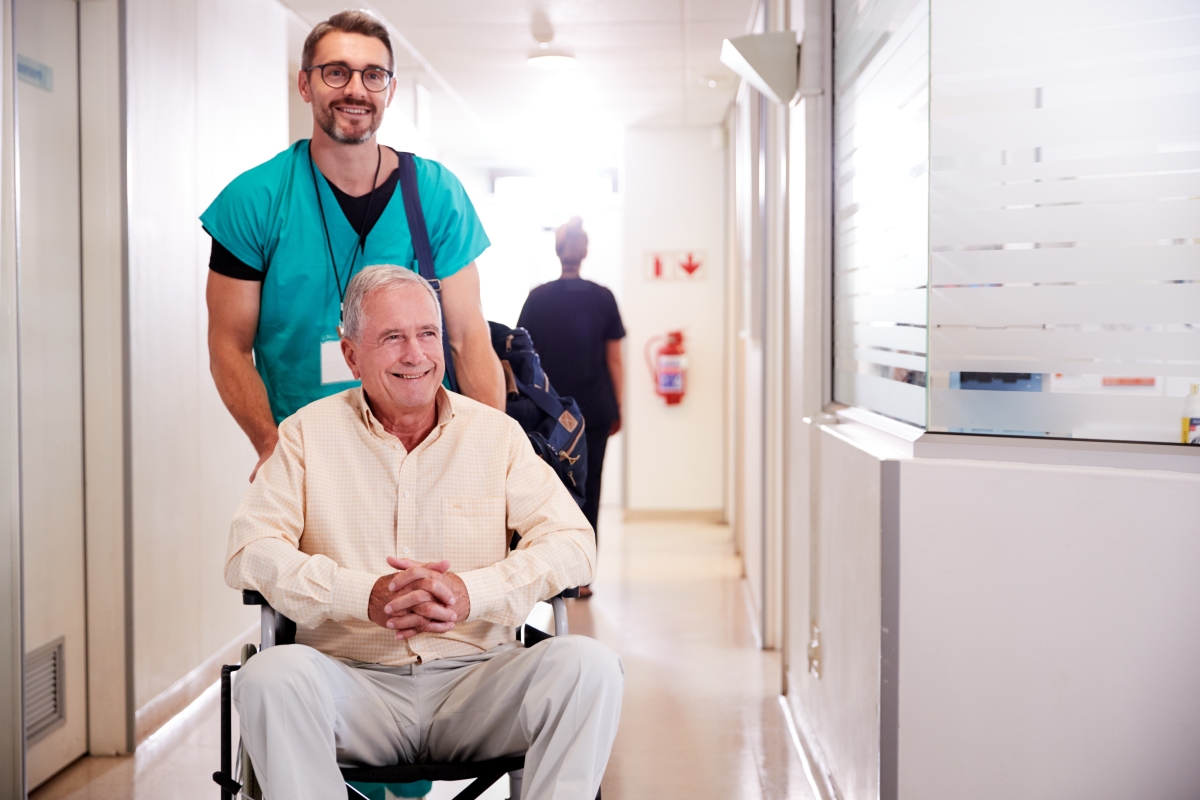 man in scrubs pushing an elderly patient in a wheelchair down a hallway