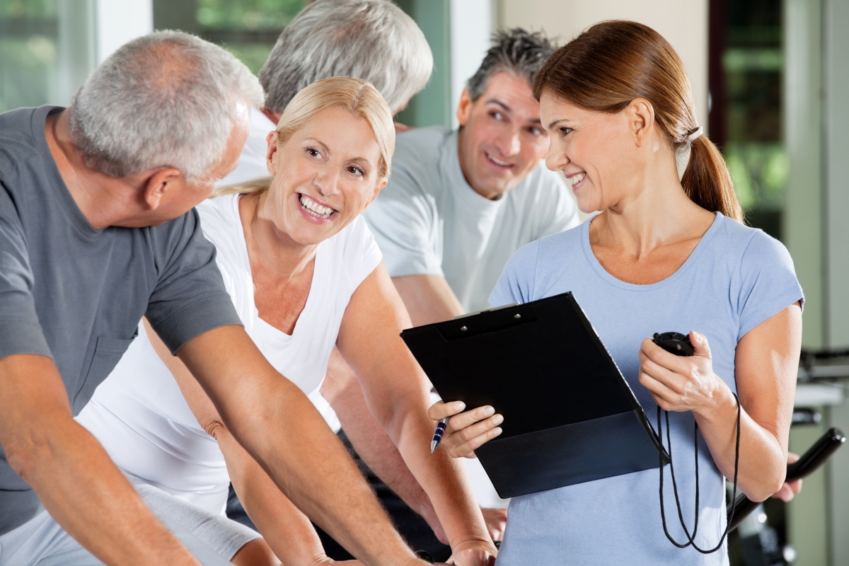 woman holding a clipboard, pen and timer while she smiles at four people indoor cycling