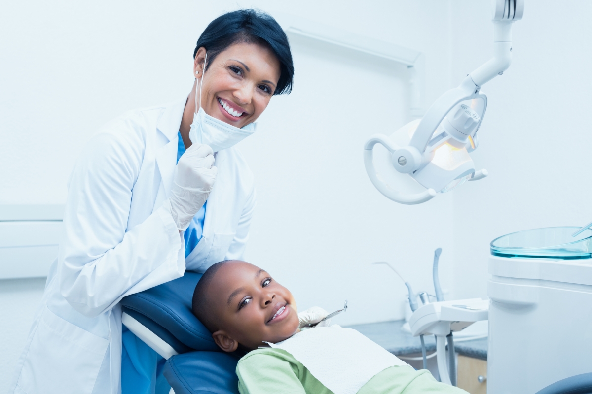 woman wearing scrubs and a mask standing next to a child reclined in dental chair with dental equipment overhead and nearby