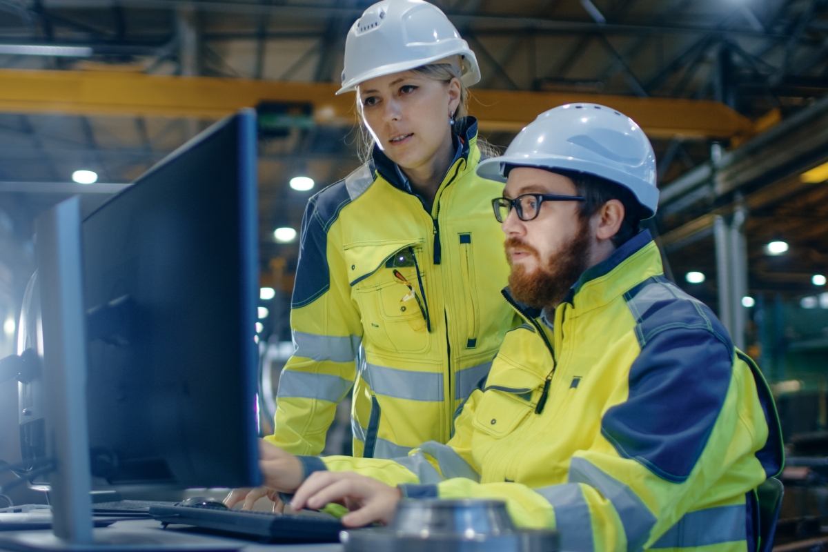two people in hard hats and safety jackets looking at a computer in a warehouse