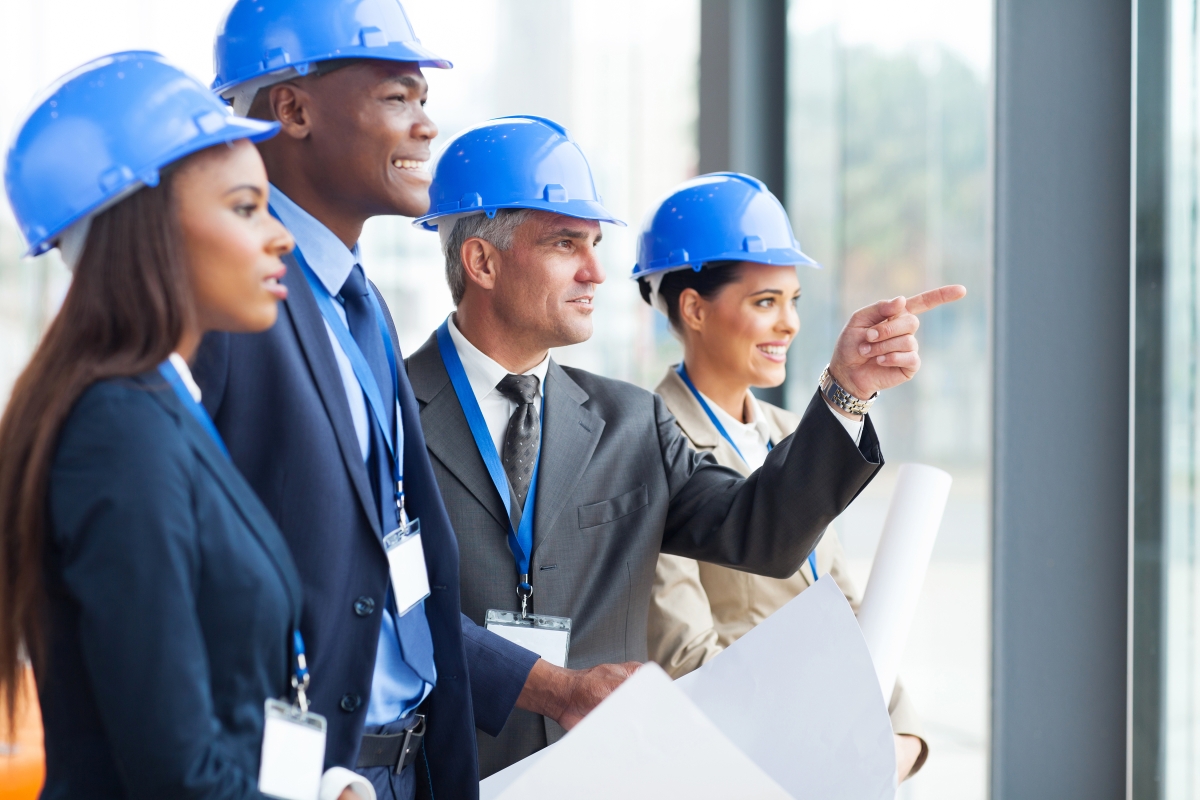 two men and two women wearing hard hats looking out a window while one of the men points out the window