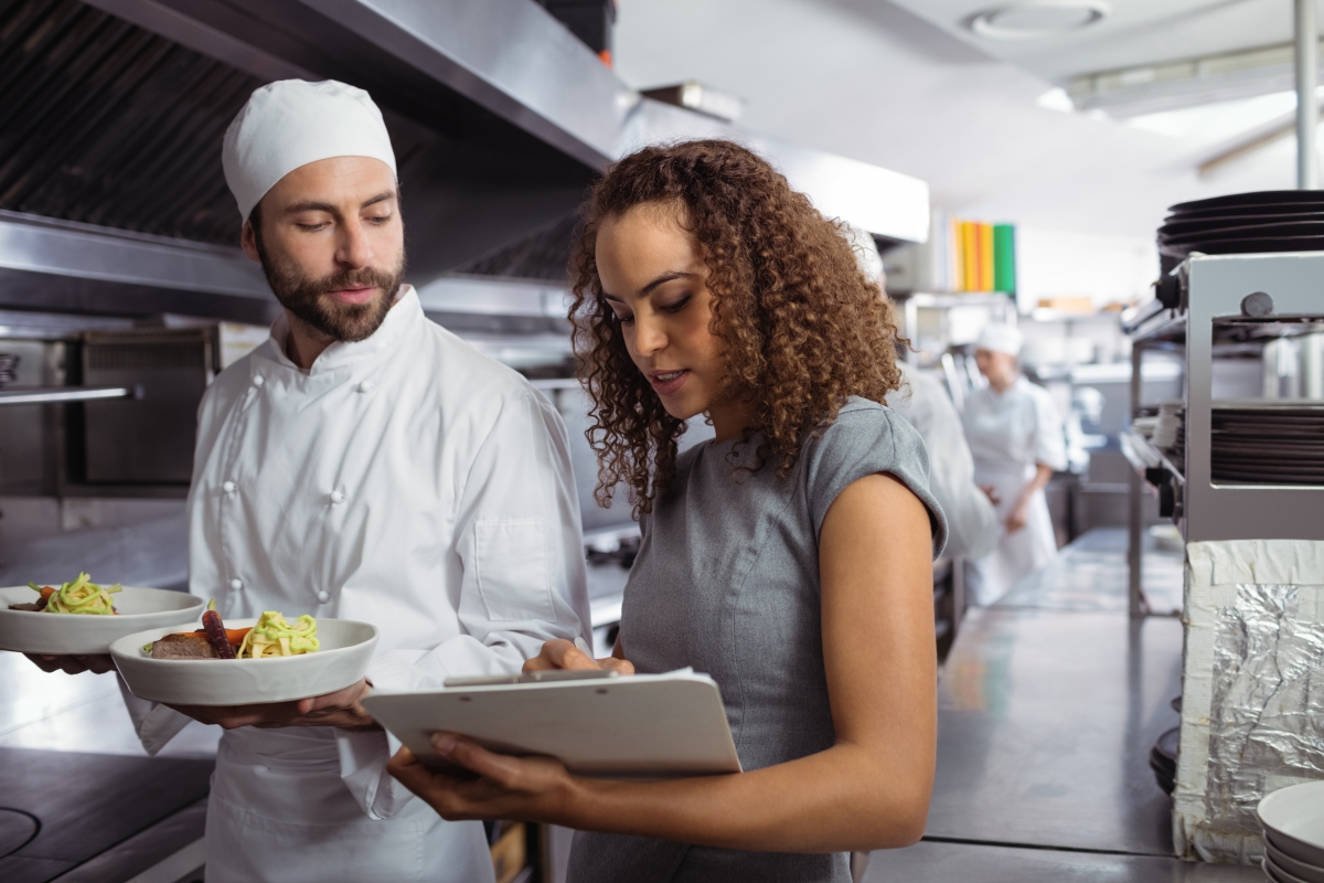 woman with a clipboard talking to a chef who is holding two plates of food in a kitchen