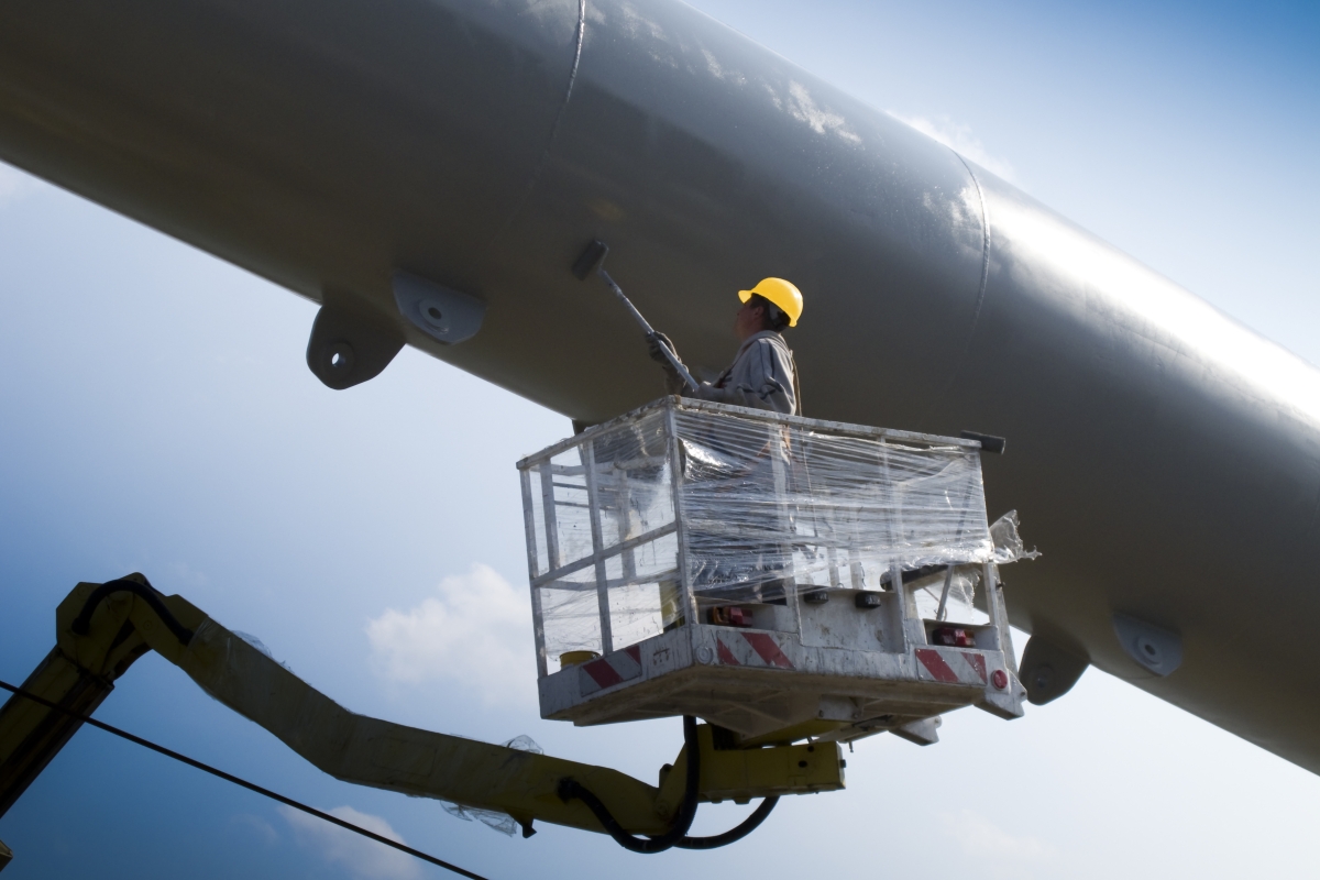 man wearing yellow hard hat painting a large structure while elevated in an aerial lift