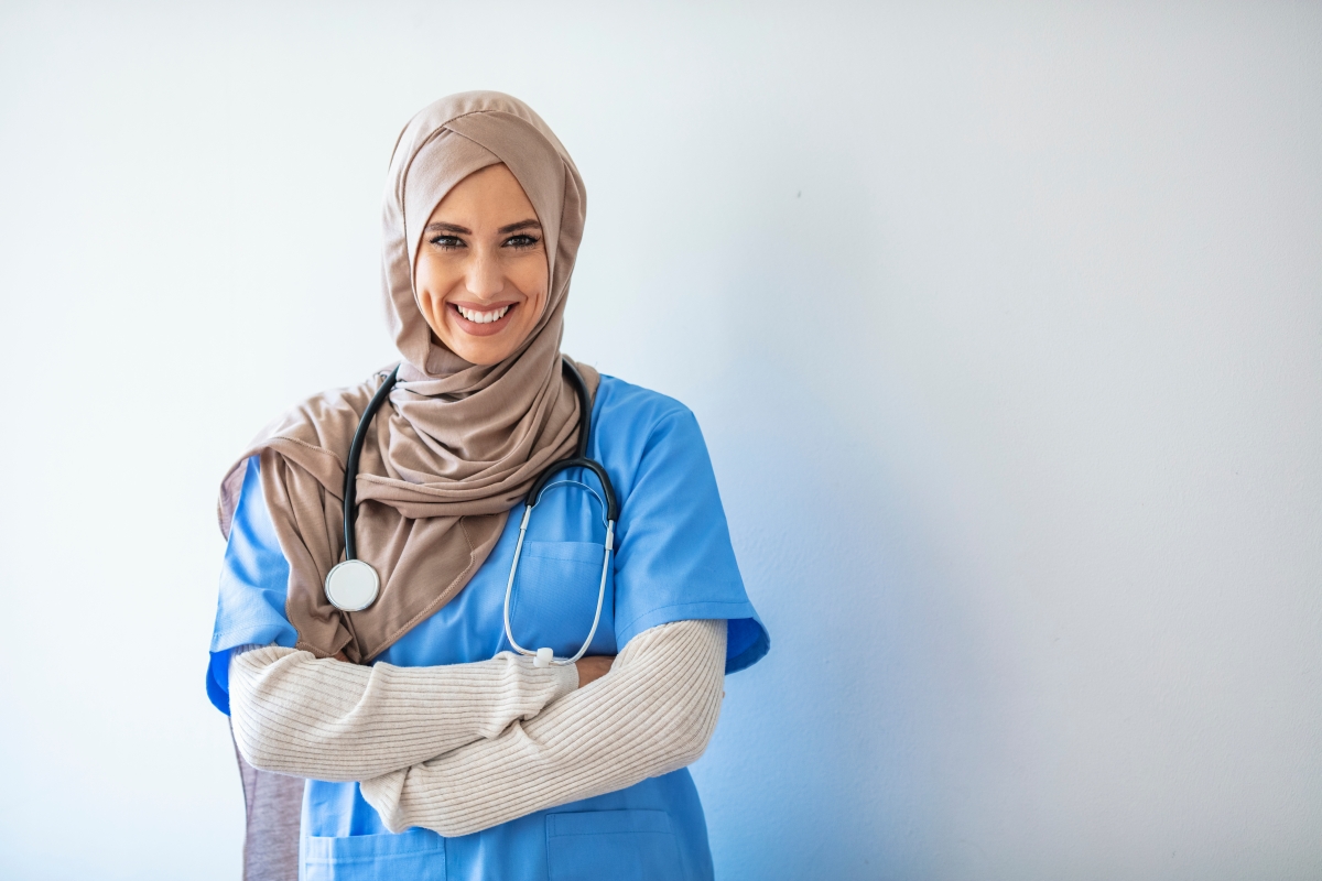 person, wearing a religious head scarf and a stethoscope, stands with their arms crossed