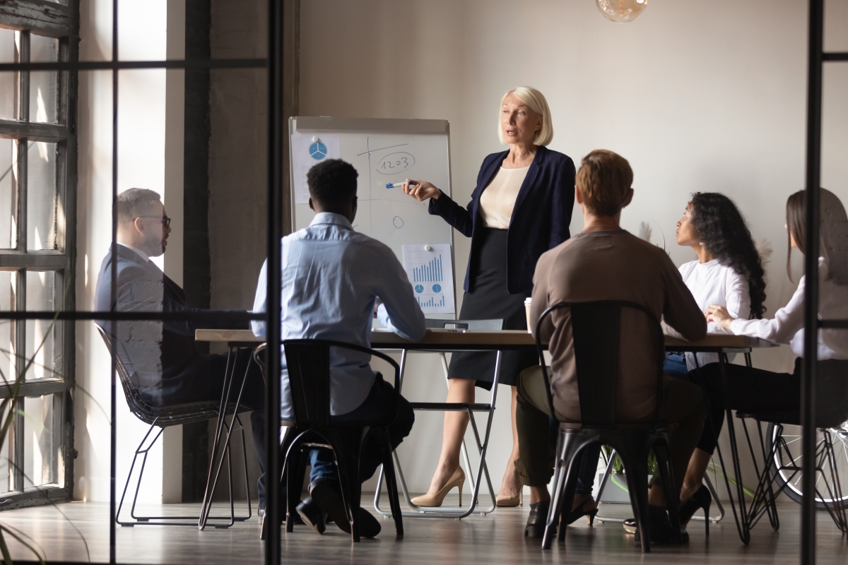 woman in business attire standing and giving a presentation to five employees seated around a table