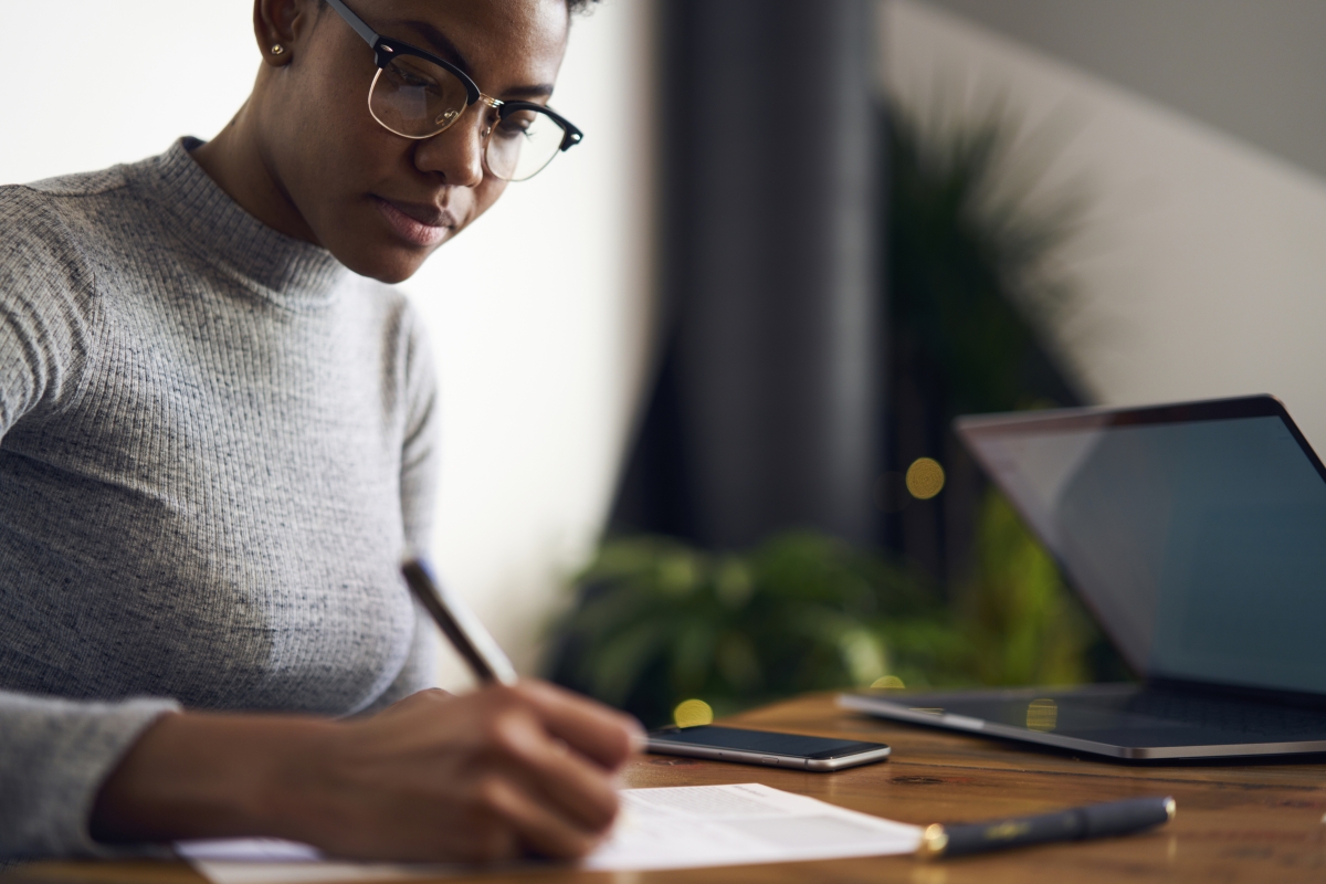woman sitting at a desk writing on paper with a laptop and smartphone next to her