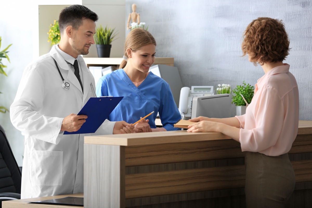 a doctor and a nurse standing behind a reception desk talking to a person on the other side of the desk while the doctor holds a clipboard and pencil