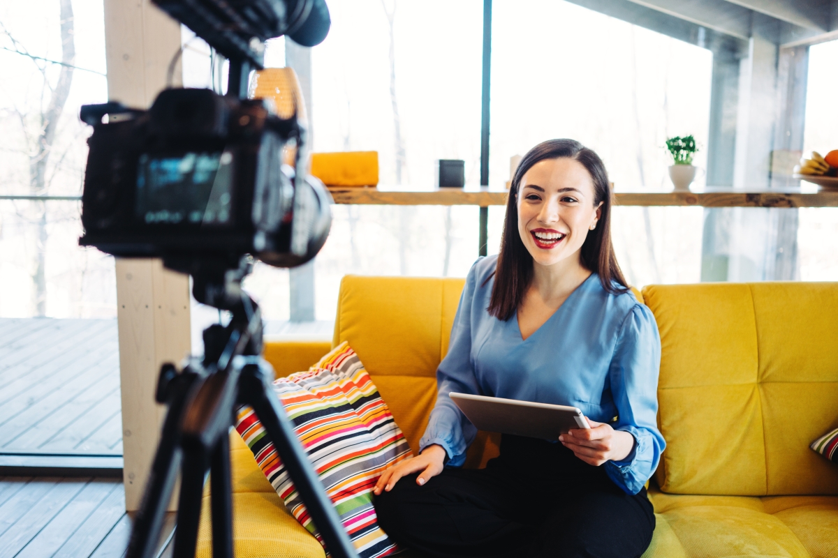 young woman sitting on a couch holding a clipboard talking to a camera and microphone set up on a tripod