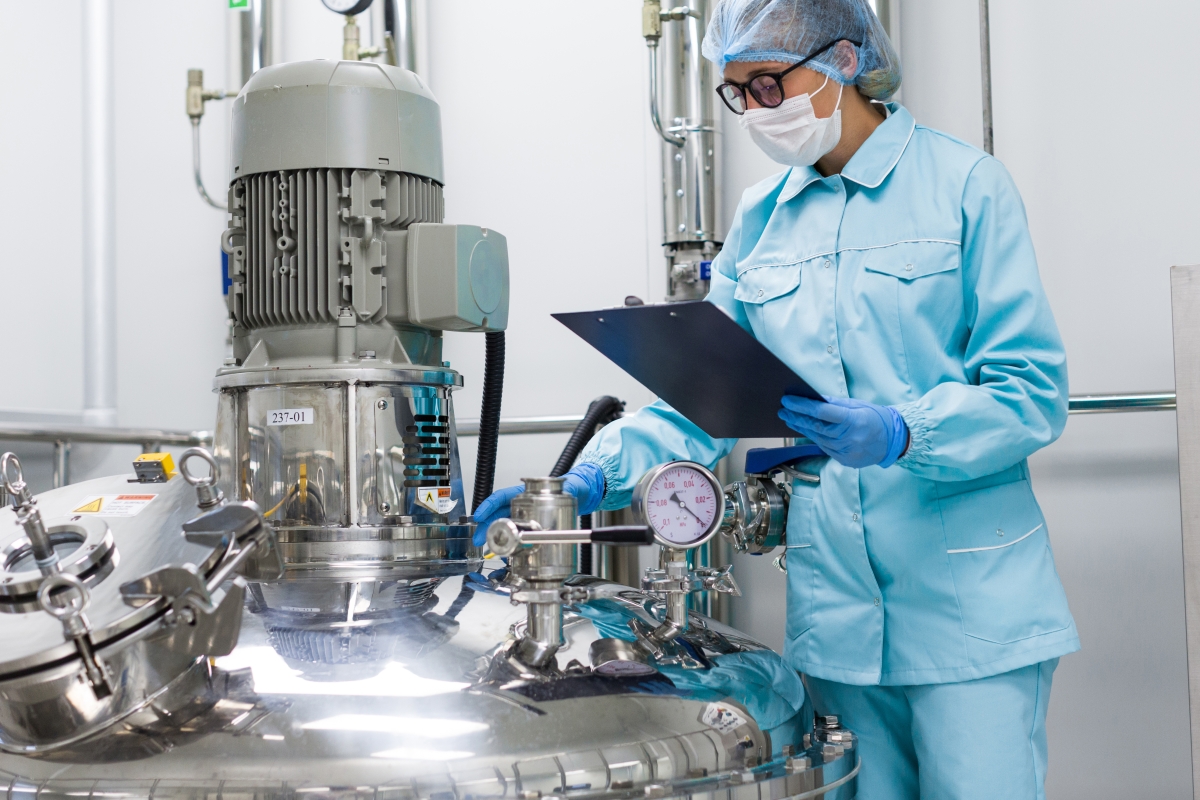 person, wearing a hair net, and mask, inspecting industrial equipment and making a note on a clip board