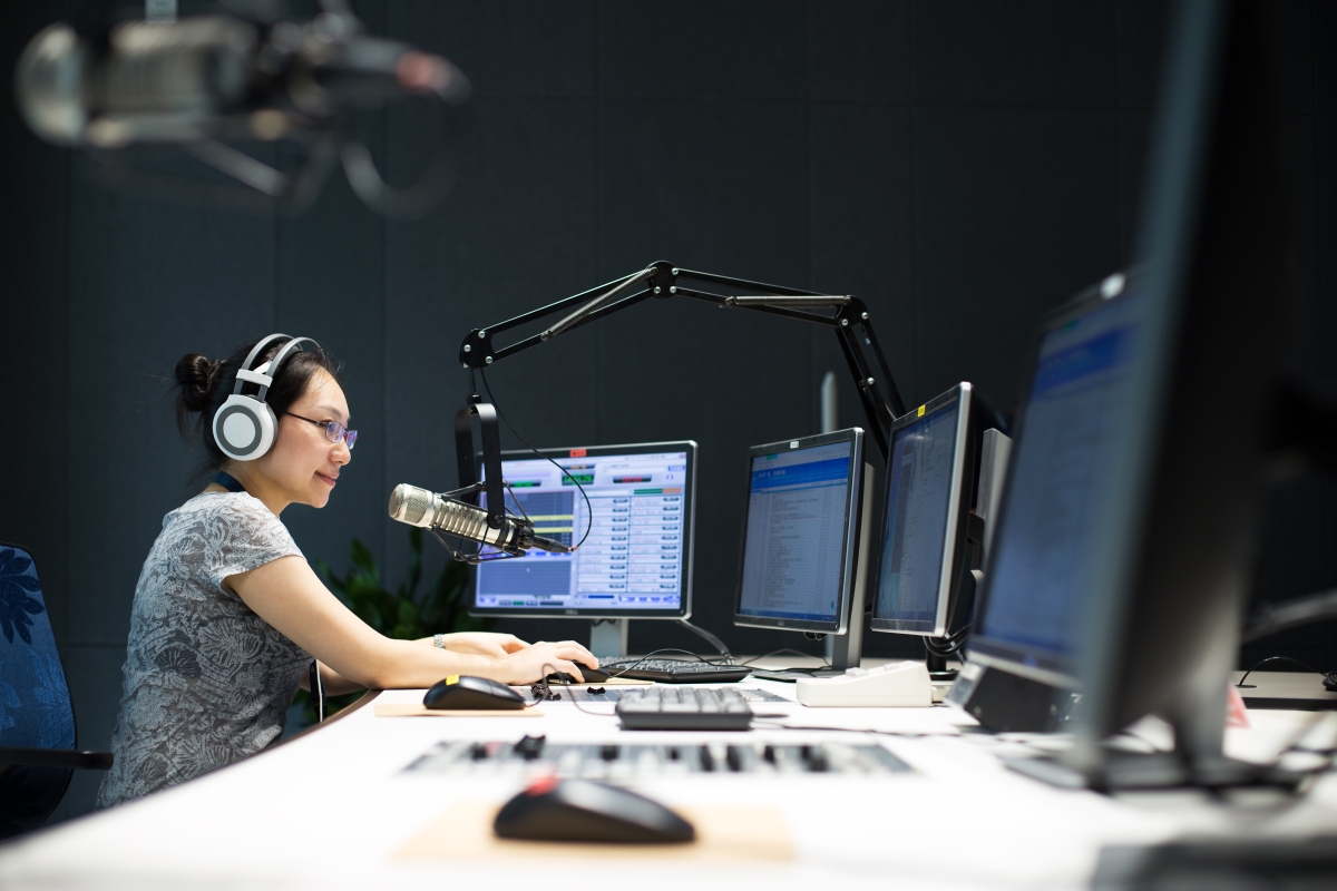 woman wearing headphones speaking into a microphone at a control desk with many computer monitors in front of her