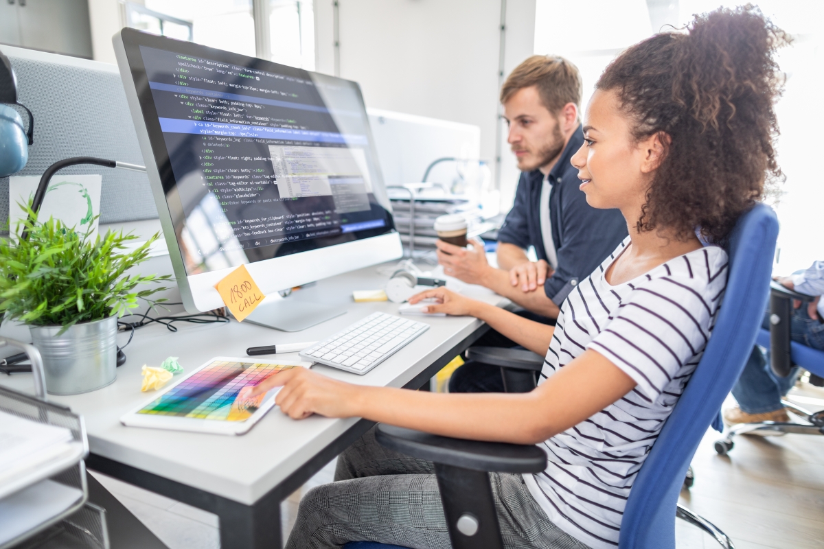 woman sitting at a desk looking at a computer displaying a screen of code while a man seated next to her looks on