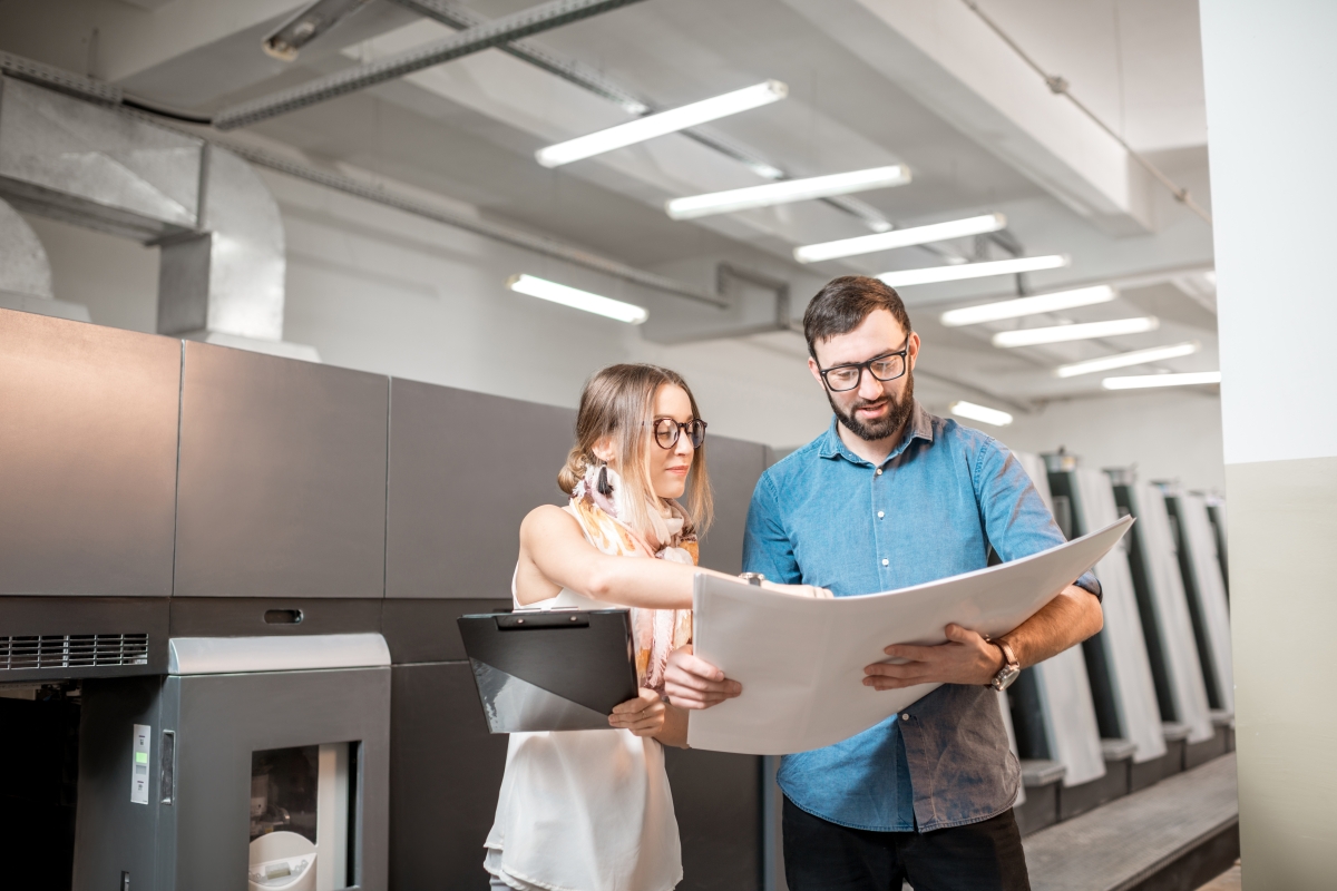 two people, standing and looking at a large piece of paper, in an industrial setting
