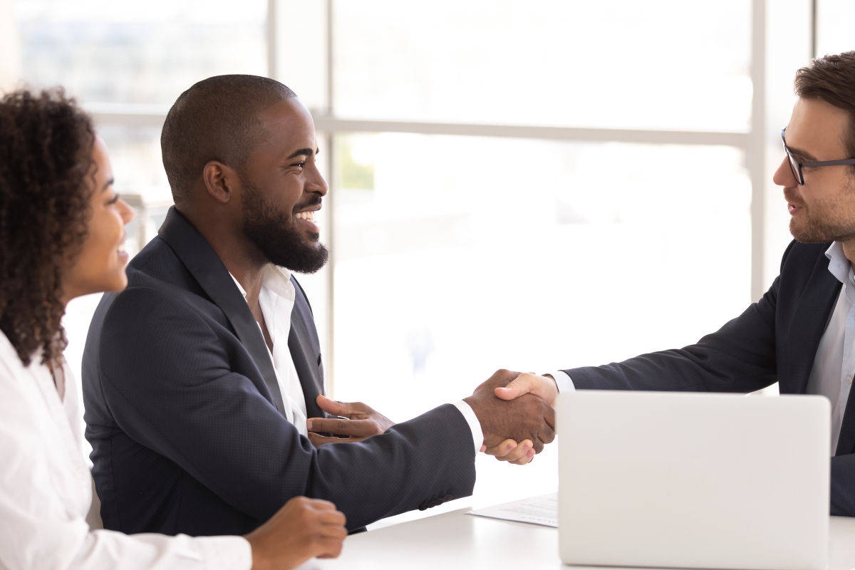 two men and one woman sitting at a table with a laptop and paper on it while the two men shake hands and everyone smiles
