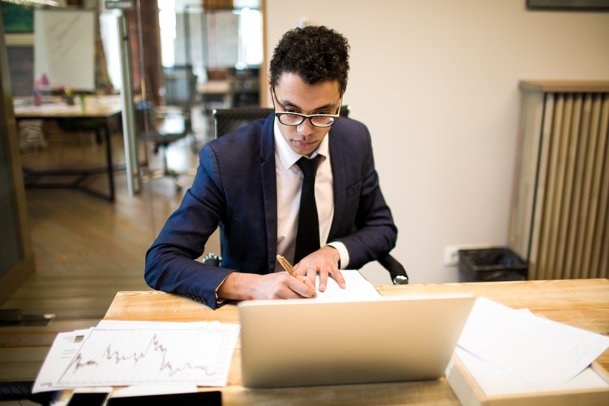man in business attire and glasses writing notes while seated at a desk with a laptop and many documents spread in front of him