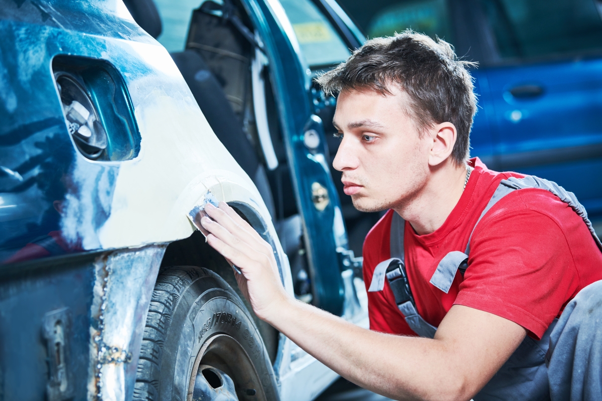 man in overalls sanding the body of a vehicle