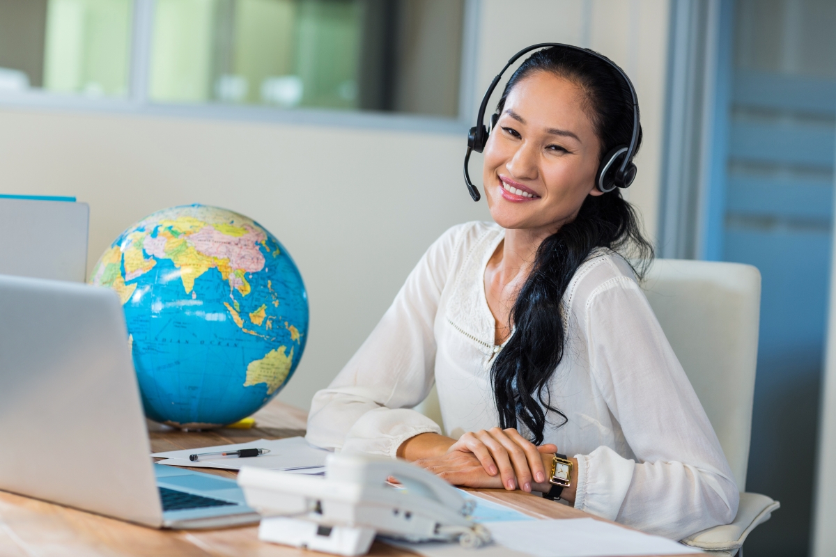 woman wearing a headset and smiling while sitting at a desk with a laptop, phone and globe on it