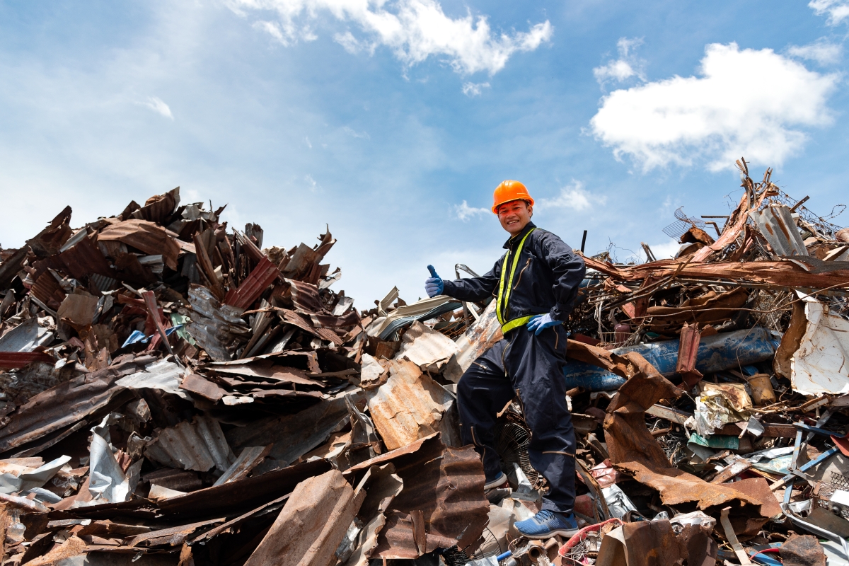 person, wearing safety equipment, standing on a pile of metal debris, giving a thumbs up