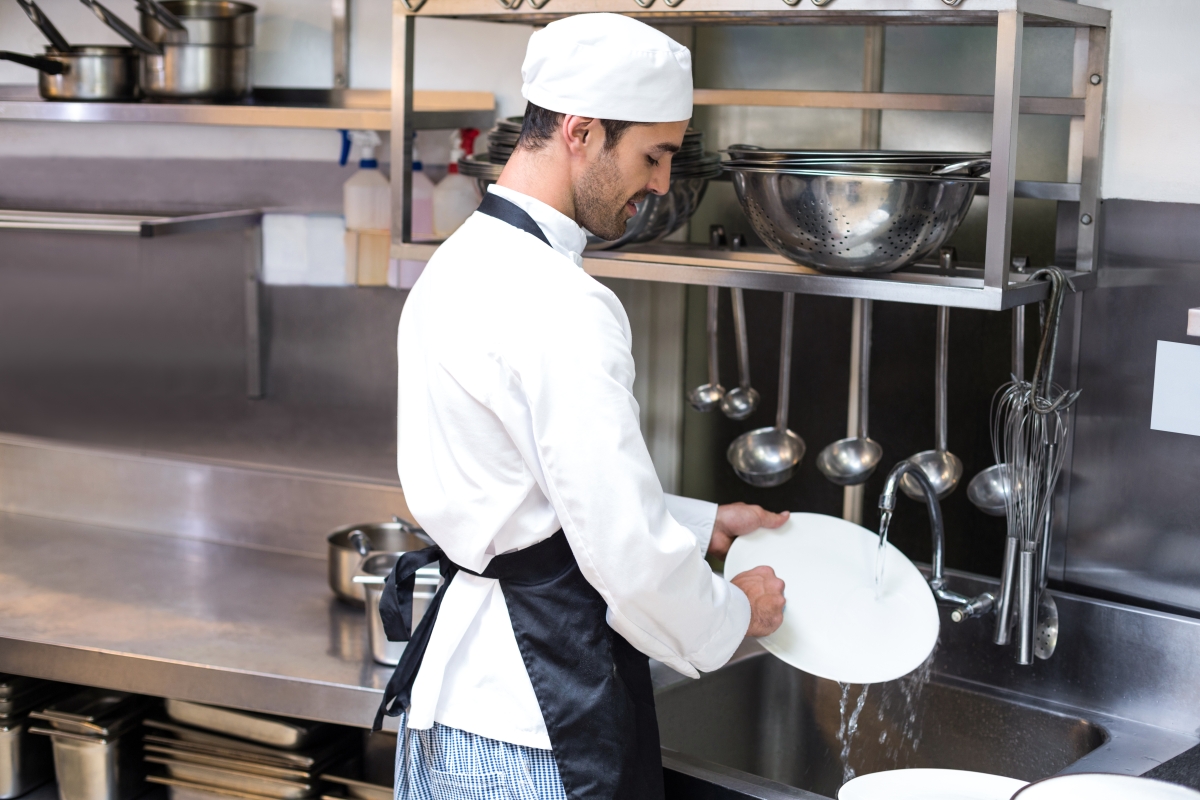 man in uniform wearing an apron and washing dishes in an industrial kitchen