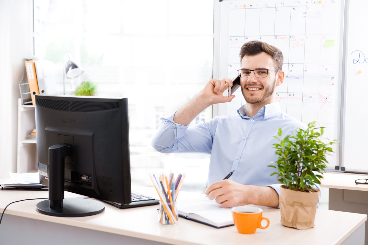 young man wearing a dress shirt and glasses sitting at a computer desk taking notes while on a phone call