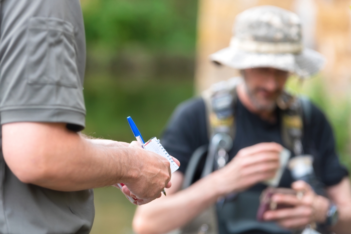 person holding notepad and pen in foreground with person in hat in background in outdoor setting