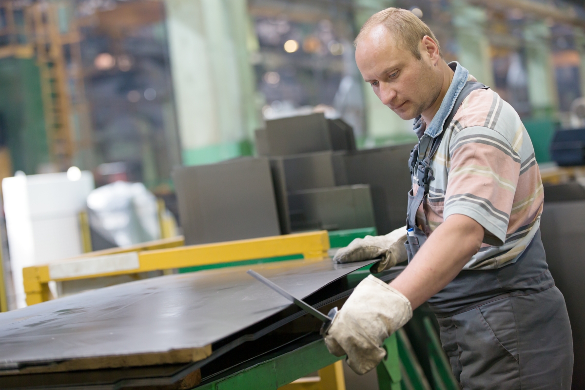 person, wearing protective gloves, measuring a piece of metal on a table in an industrial setting