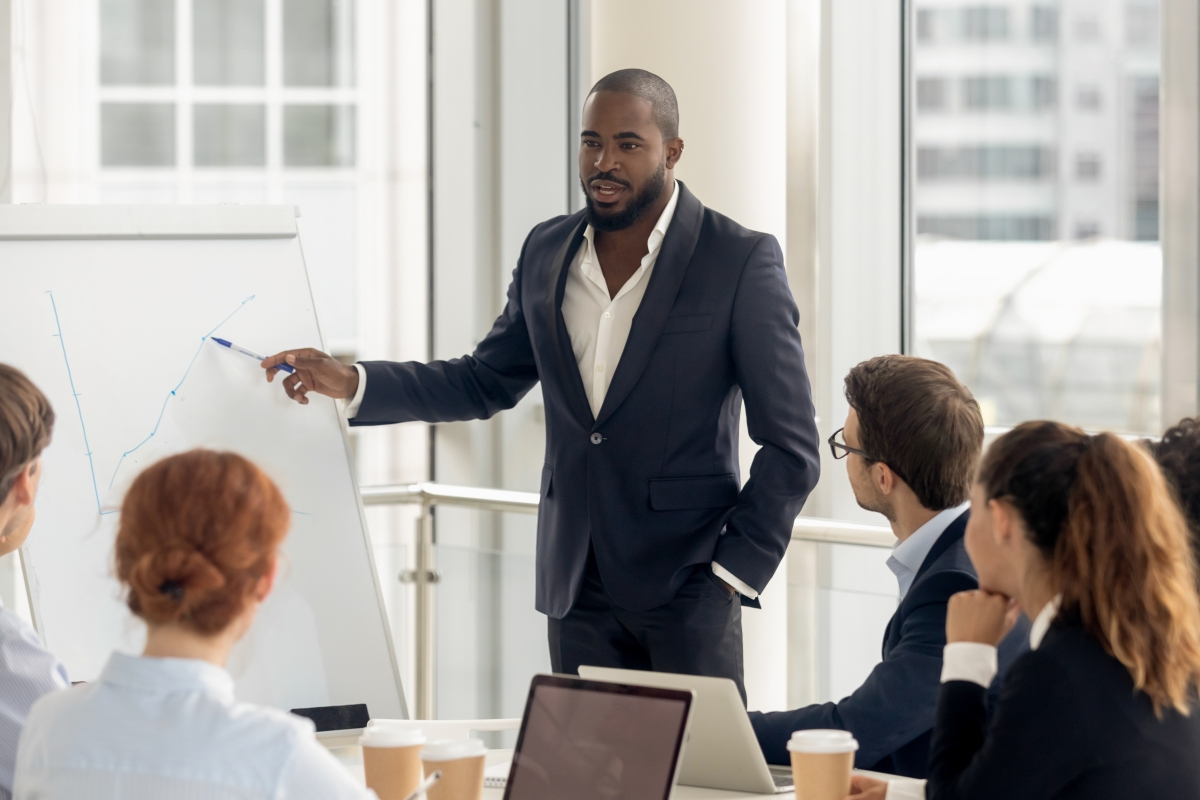 man in professional business attire standing and using a pen to point at a graph on a board while giving a presentation to a group seated around a table