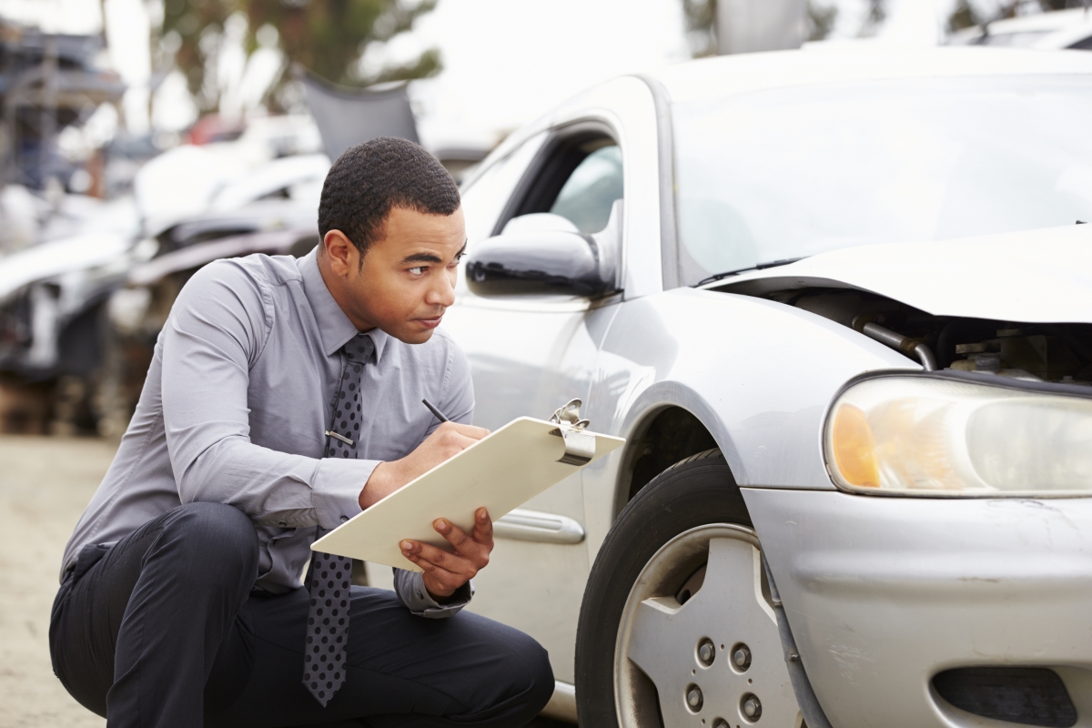 man in professional attire inspecting a damaged car while crouched down next to it writing on a clipboard