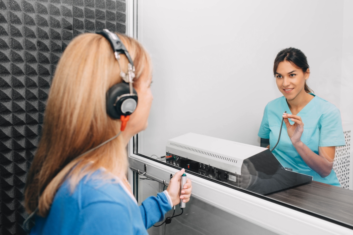 woman in scrubs seated at a table with a device in front of her assessing a client wearing a headset sitting across from her with a screen between them
