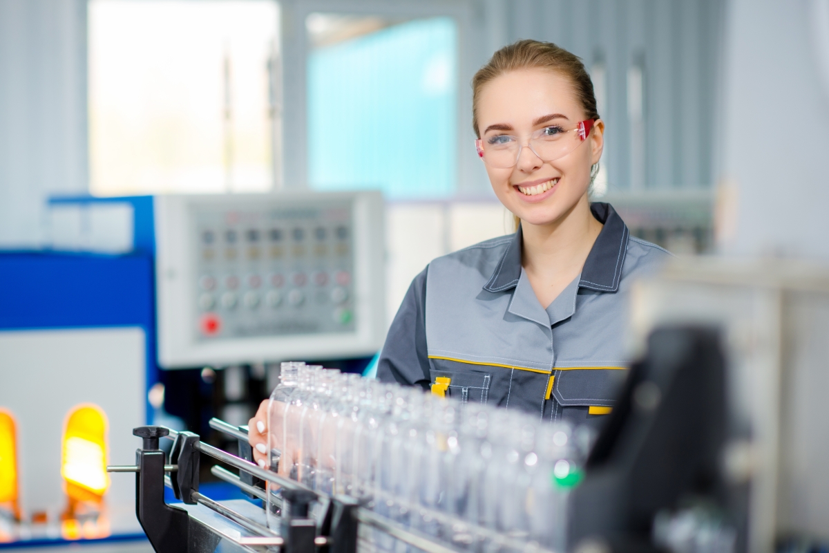 person, wearing safety goggles, standing near a row of plastic bottles on an assembly line
