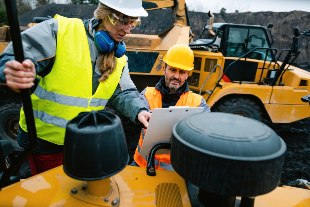 man and woman wearing safety vests and hard hats looking at a clipboard while on construction or mining site surrounded by large machinery and equipment