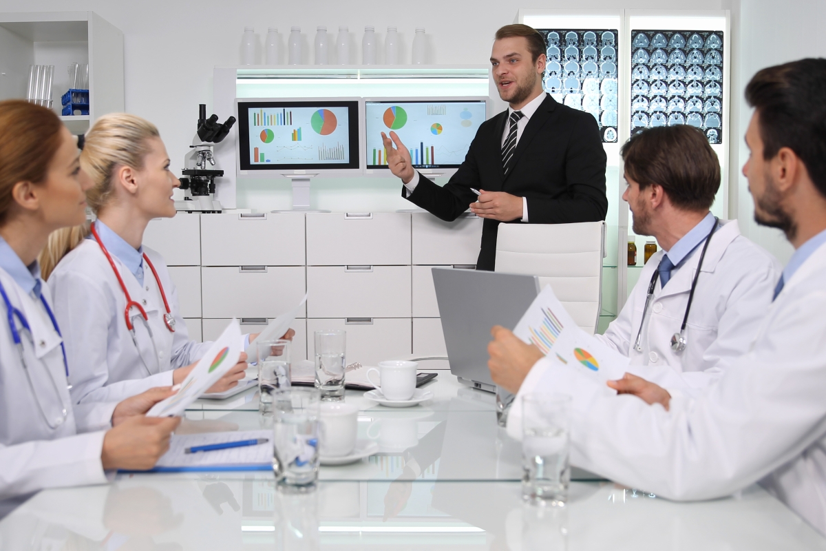 man in a suit standing and giving a presentation to two female and two male doctors that are all seated at a table in a medical office setting